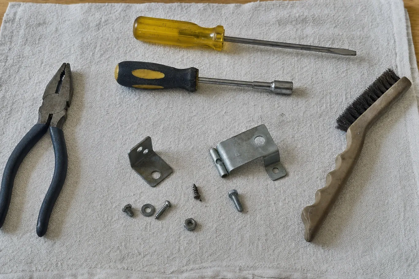 Pliers, a flat driver, a nut driver, two sheet-metal brackets, loose fasteners and a wire brush on a folded cloth
